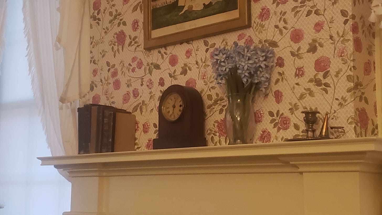 The fireplace mantle in Dickinson's bedroom with a set of small books, a small rounded mantle clock showing 6:04. A vase of purple star-like flowers, and a brass candlestick with snuffer. The edge of a small framed painting is above the mantle. 