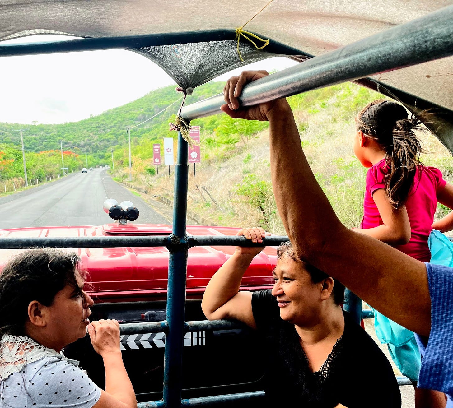 Two women in the back of a truck talk as a little girl stands up looking ahead as the truck drives through a green valley