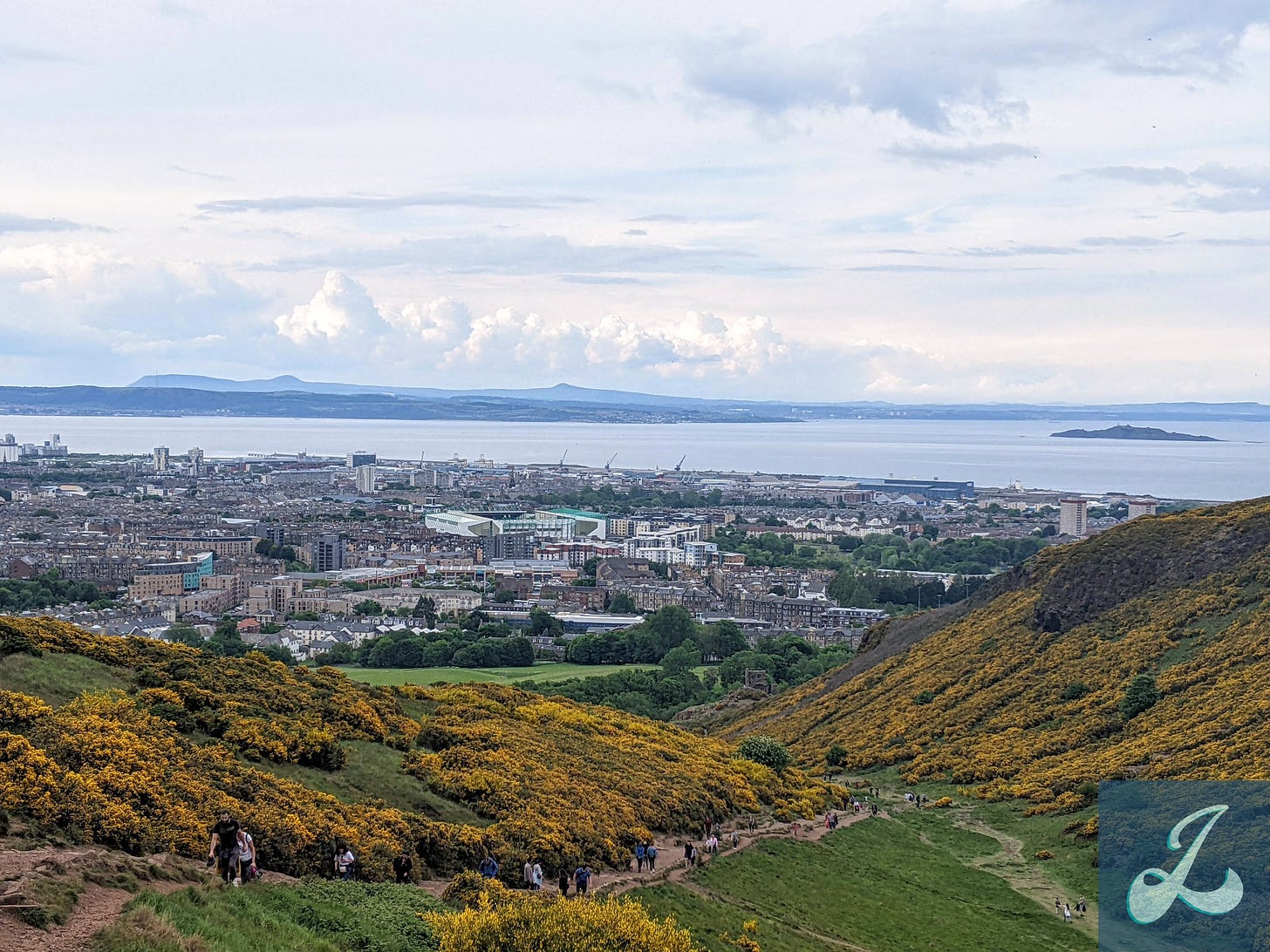A landscape photo of Edinburgh. There is a moderately cloudy sky above the bay and cityscape of Old Town Edinburgh. In the foreground is a rocky dirt path leading to the city, surrounded by hills covered in thick patches of yellow, spiny flowers called gorse. Several people are walking up the path away from the city.