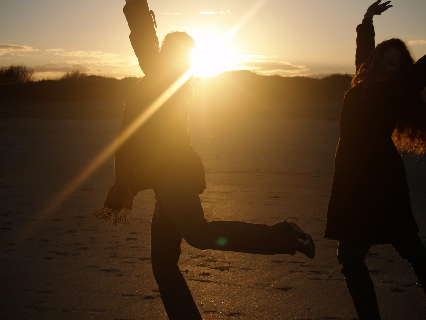 Two women silhouetted against a setting sun, dancing and twirling on a beach. 