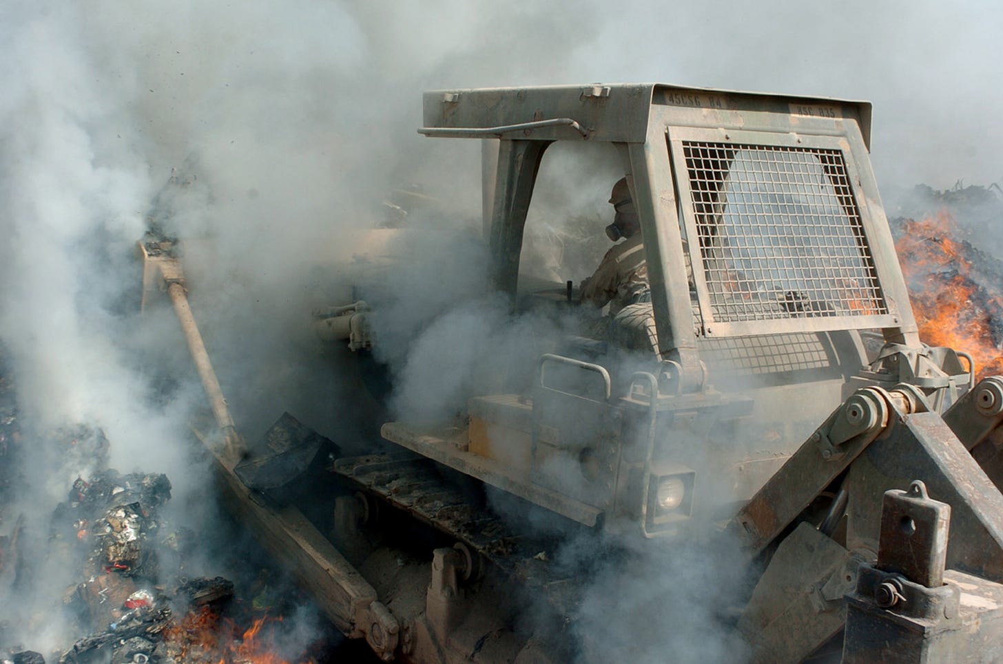 Soldier operates bulldozer in Iraq burn pit, enveloped in smoke.
