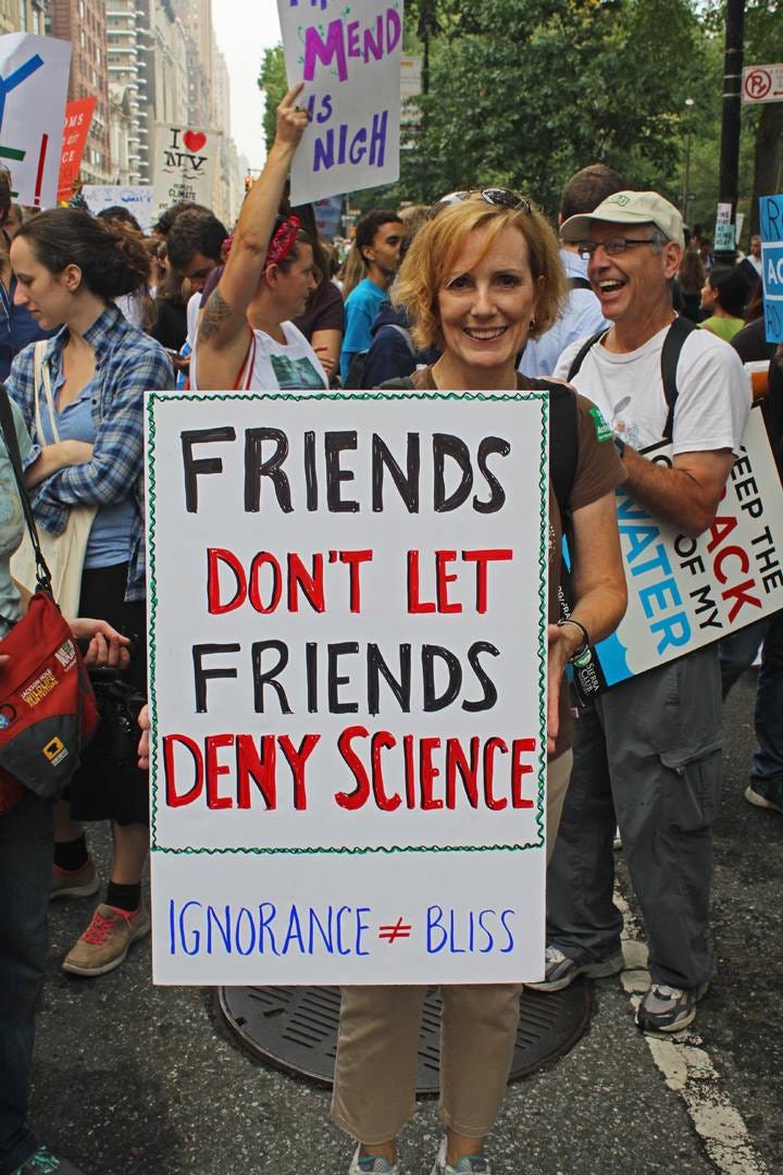 People at a protest with climate-related signs. A woman is smiling and holding a sign that says, "Friends don't let friends deny science. Ignorance does not equal bliss."