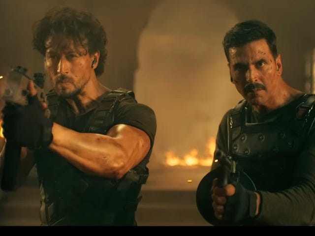 A still from Hindi film, ‘Bade Miyan Chote Miyan’. Freddy (Akshay Kumar) and Rocky (Tiger Shroff) in combat overalls pointing their guns towards the camera in a yellowish brown, ablaze background. 