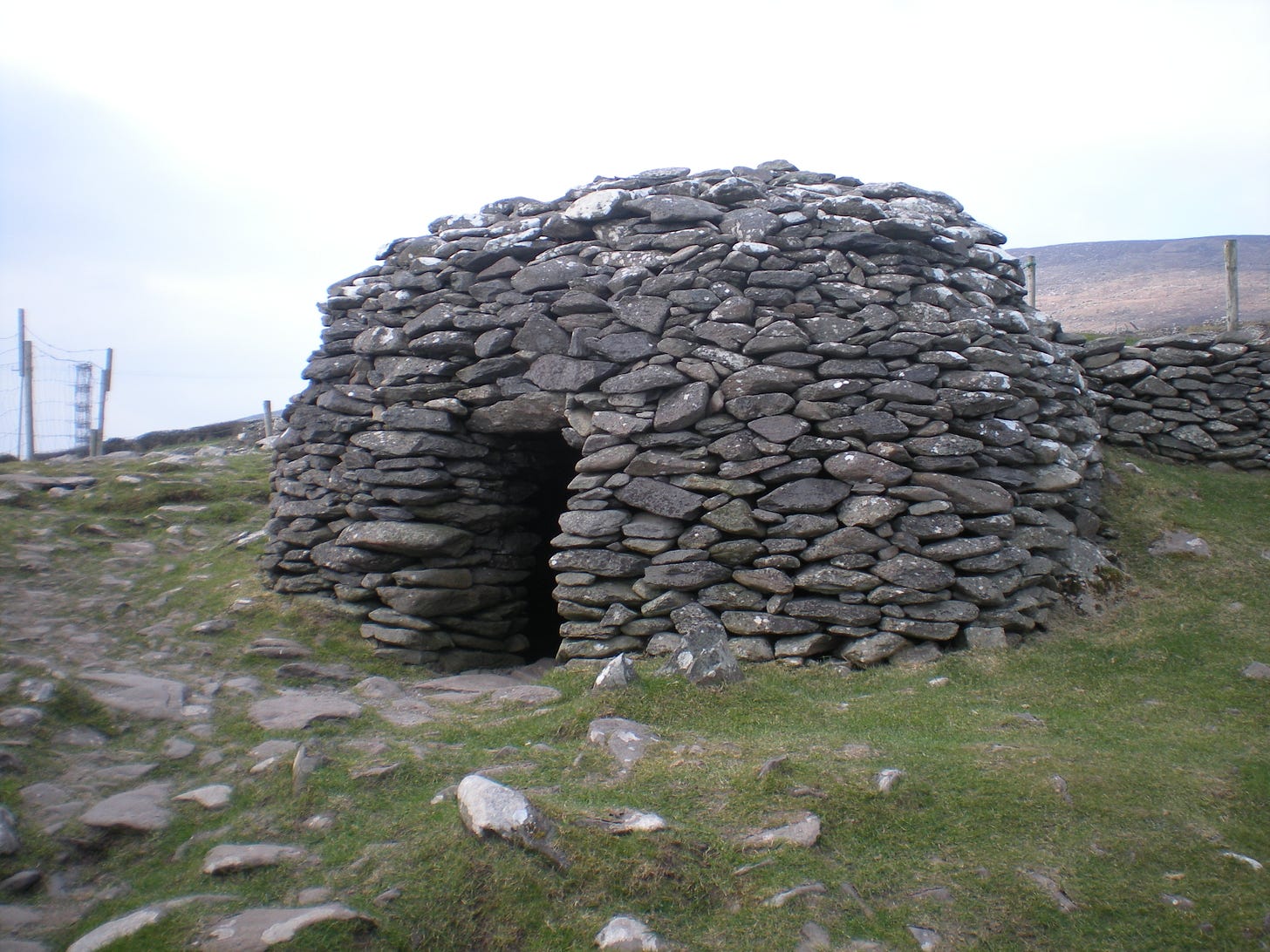A round, hut with a low open doorway sits on a green hillside with other rocks strewn around.