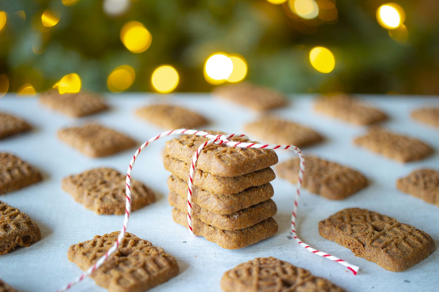 Patterned, rectangular golden biscuits are placed on a white table, with some balanced in a tower and ties with red and white festive bakers' twine. Behind are bokeh fairy lights.