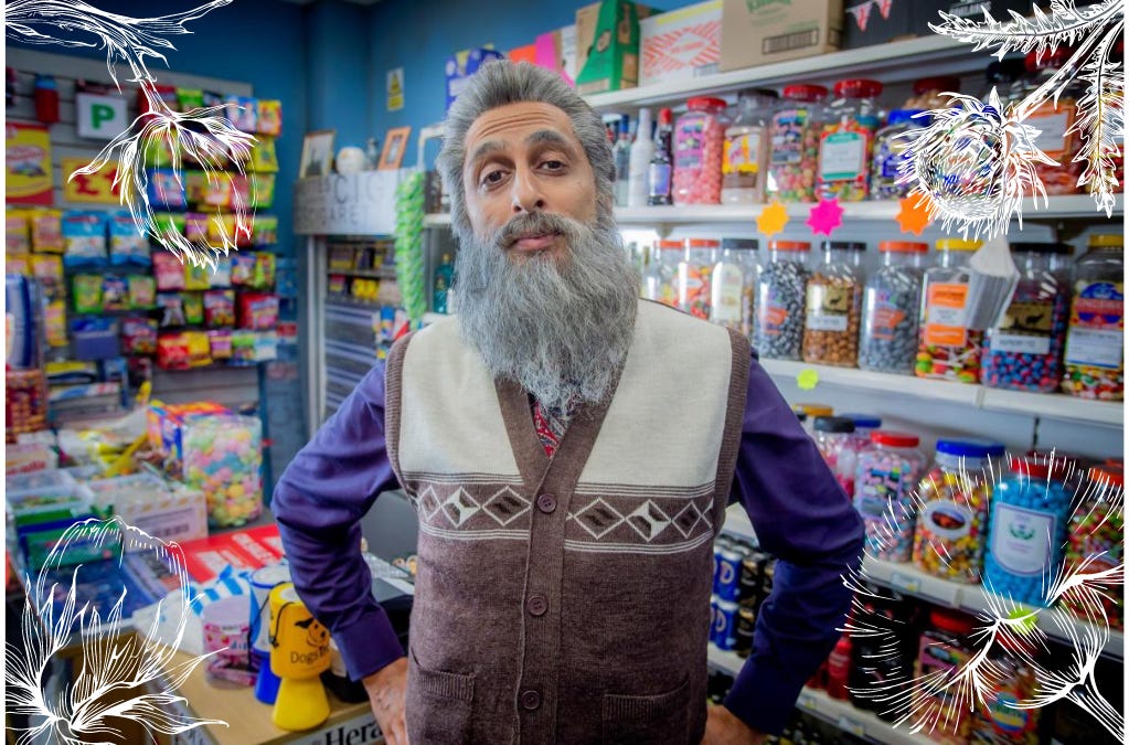 Elderly Navid Harrid (played by Sanjeev Kholi) stands facing the camera with his arms resting on his hips. He is in his corner ship, and we can see rows of sweet jars behind him. To his right is the shop counter, with a till, more sweets, a few charity tins and some boxes of chewing gum sitting on top.