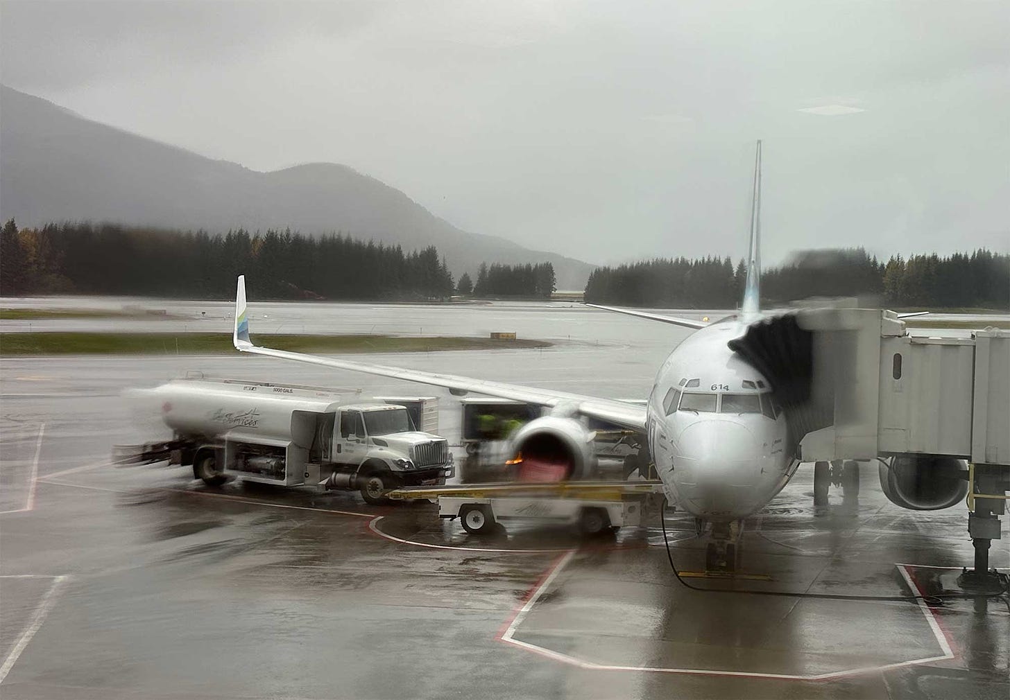 Alaska Airlines Boeing 737 parked at gate in Juneau, seen through rain-soaked and blurry window.