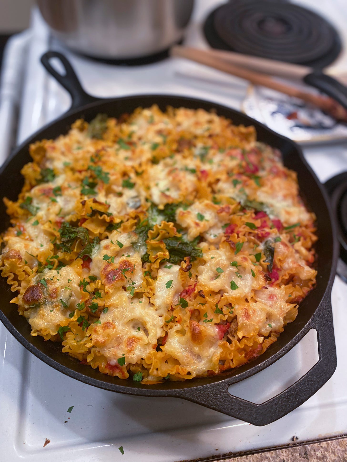 A large cast iron pan filled to the brim with baked pasta in a light tomato sauce with kale and basil, topped with patches of mozzarella and dusted with parsley.