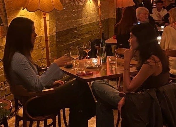 This may contain: two women sitting at a table in a restaurant talking to each other and drinking wine