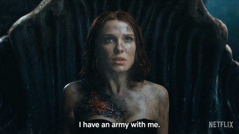 a gif of Elodie from the movie 'Damsel', played by Millie Bobby Brown. She is saying "I have an army with me" while breathing heavily, with a dark background behind her, and a burn on her left shoulder.