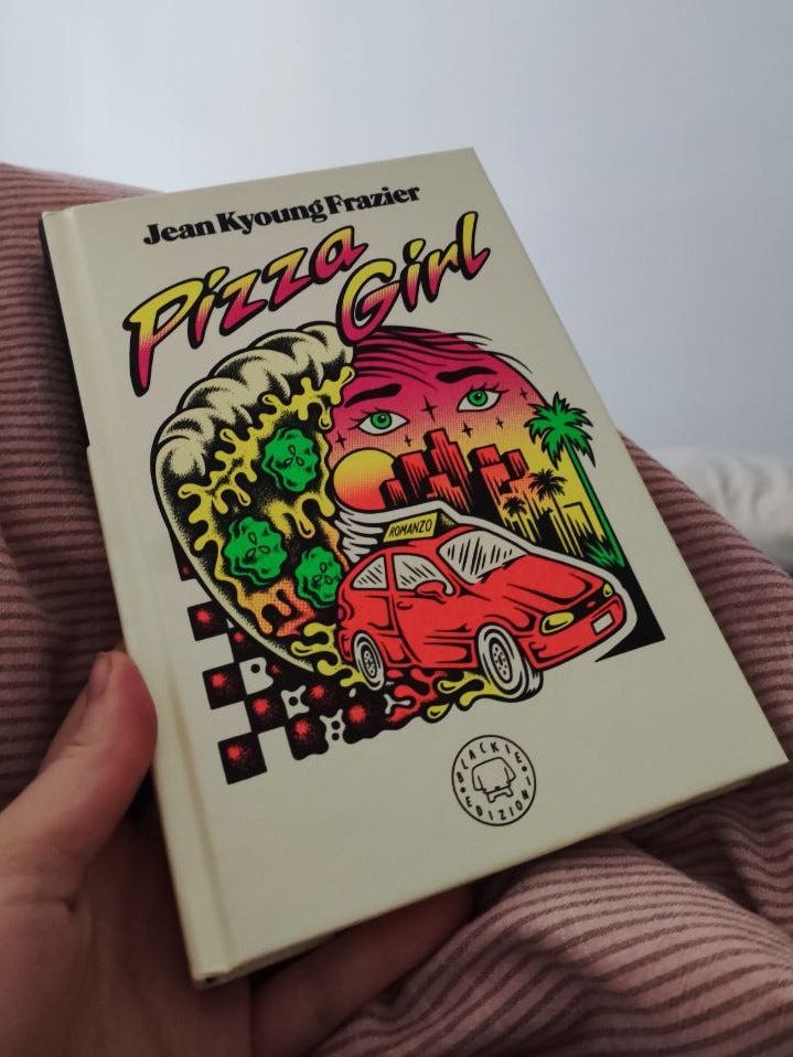 Una mano tiene un libro. Sullo sfondo un letto e le coperte. Il libro è Pizza Girl. C'è un'illustrazione molto colorata con una macchina che sfreccia, una fetta di pizza, degli occhie  una città