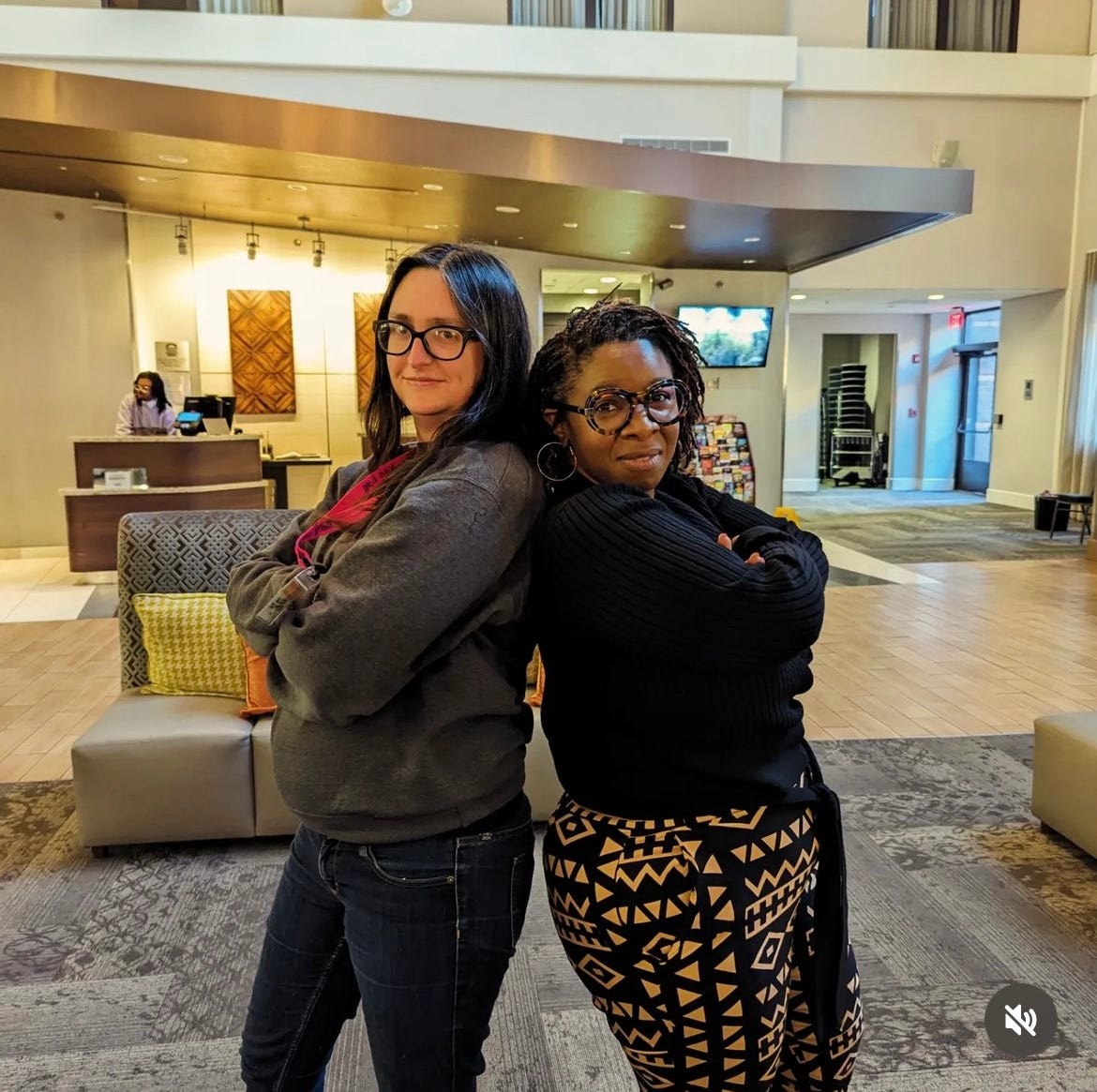 Picture of me, a white woman wearing black glasses and a gray sweatshirt, back-to-back with Nikki Payne, a Black woman wearing circular tortoiseshell glasses and geometric patterned pants