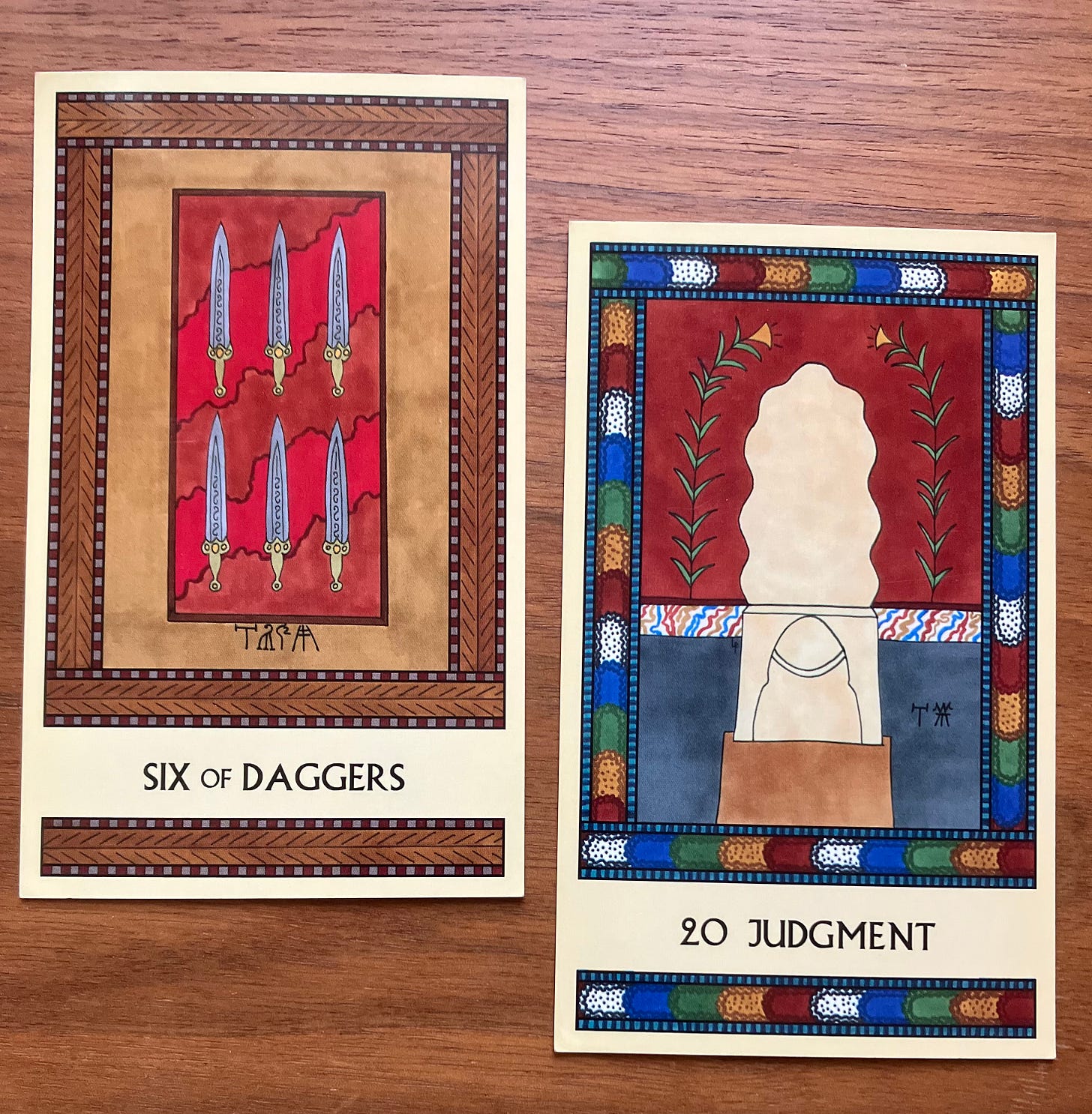 Two Minoan Tarot cards on a brown wood surface. The Six of Daggers is in shades of tan, brown, and red. It shows six fancy daggers lying in a decorative presentation box. Judgment has a multicolored border. It shows the central seat in the Knossos Throne Room, surrounded by painted walls.