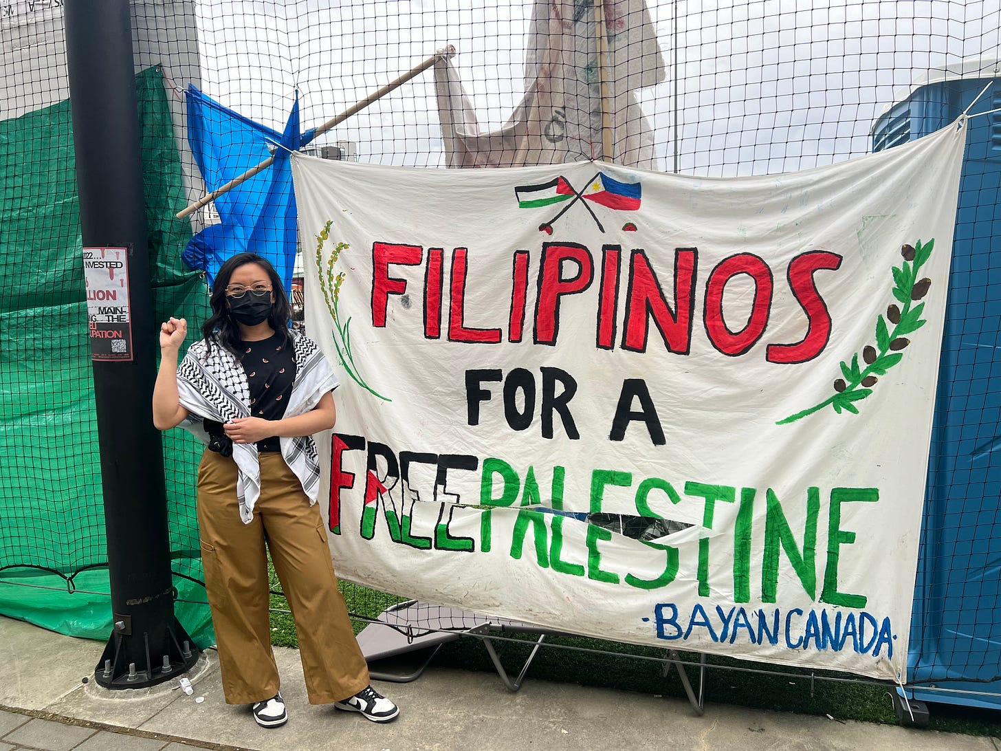 Author Justine Yu, wearing brown pants, a black patterned shirt, and a keffiyeh, stands next to a large Filipinos for a free Palestine banner, written in red, black, and green.