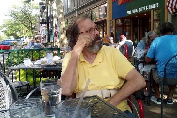 David Shaw sits at an outdoor table, looking off to his left while buckled into his wheelchair.