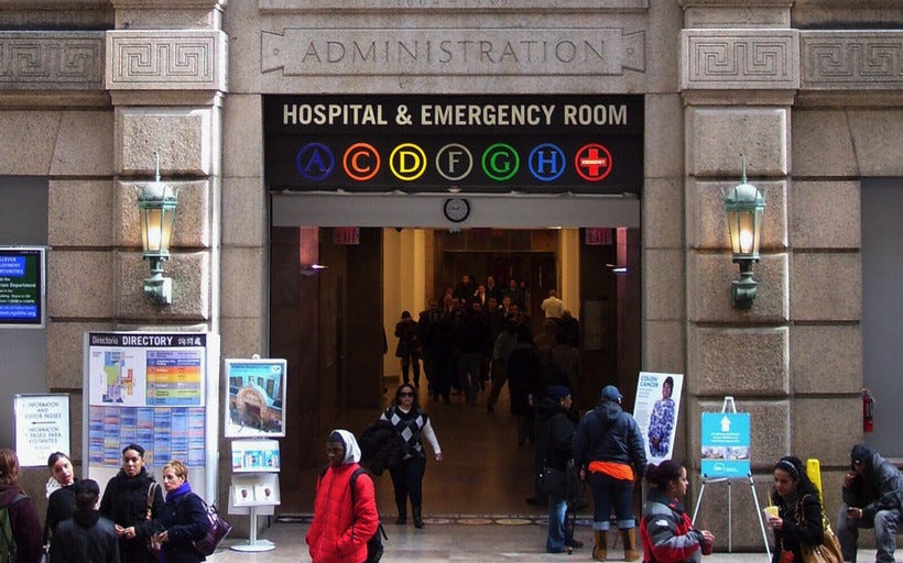 People gather outside a stone arch with sign that says "Hospital & Emergency Room" displaying letters A, C, D, F, G, H, and an emergency cross.