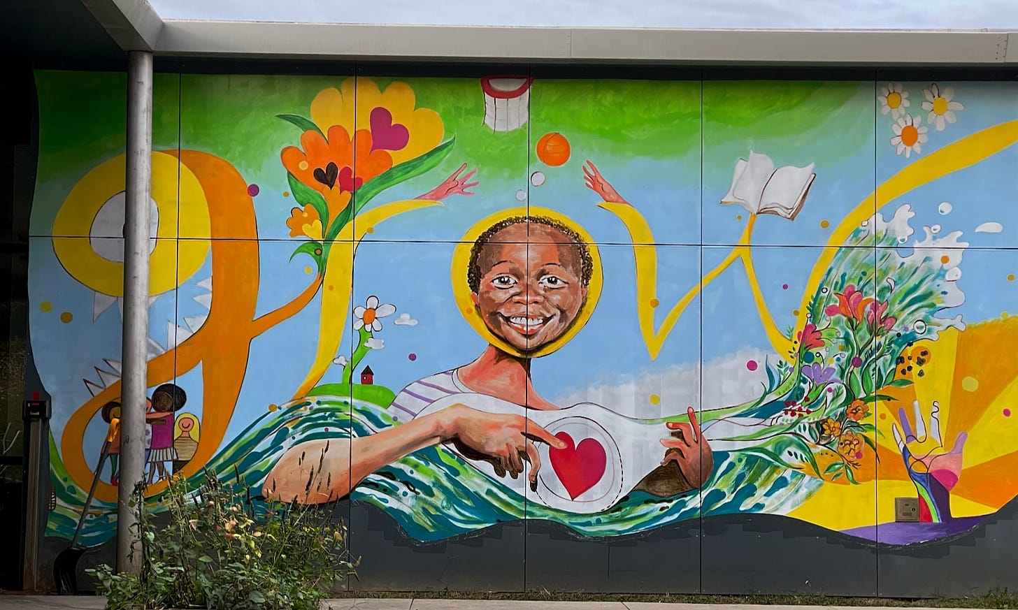 Mural with the word grow that shows a Black boy with his smiling face within the small “o”. He is holding an object in a way similar to how a guitar is typically held. There is a heart in the middle and a bouquet of flowers at the end. Other elements includes children interact, a green wave, a basketball, a book, a hand with sunshine coming out of it, and flowers.