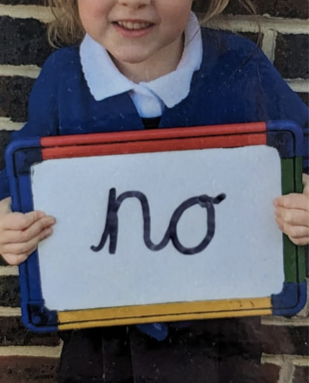 Hayley's five-year-old daughter proudly holds a sign that bears the word 'no' in her own handwriting. She is wearing a school uniform and stood against a brick wall. Her face isn't visible but her broad grin is.