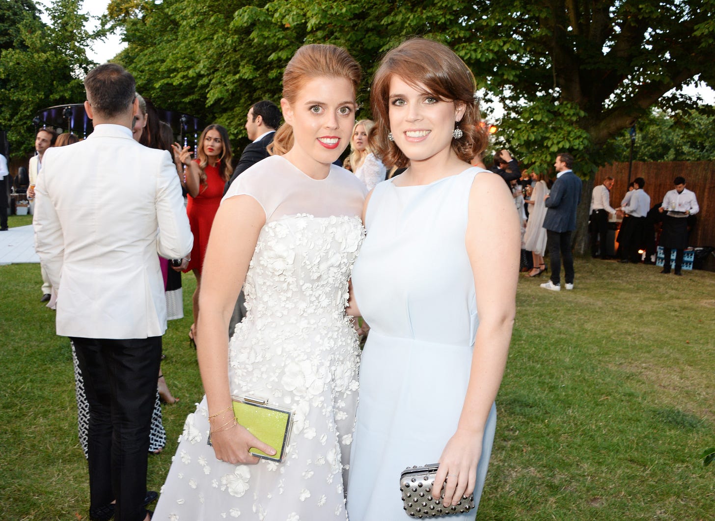Princess Eugenie on the left in a white dress and Princess Beatrice on the right in a blue dress, both smiling