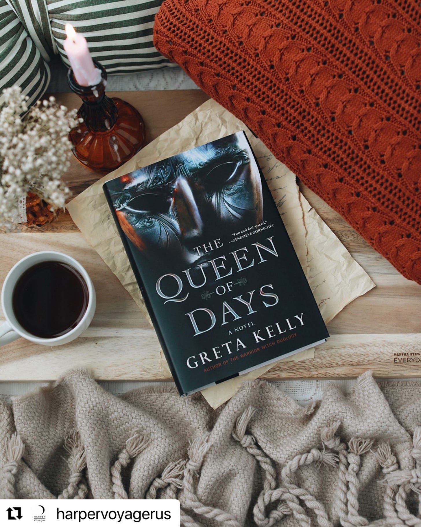 A picture of a black book with a silver mask on the cover and the title The Queen of Days. The Book is sitting on a wooden table beside a cup of coffee, a candle and brown and orange blankets