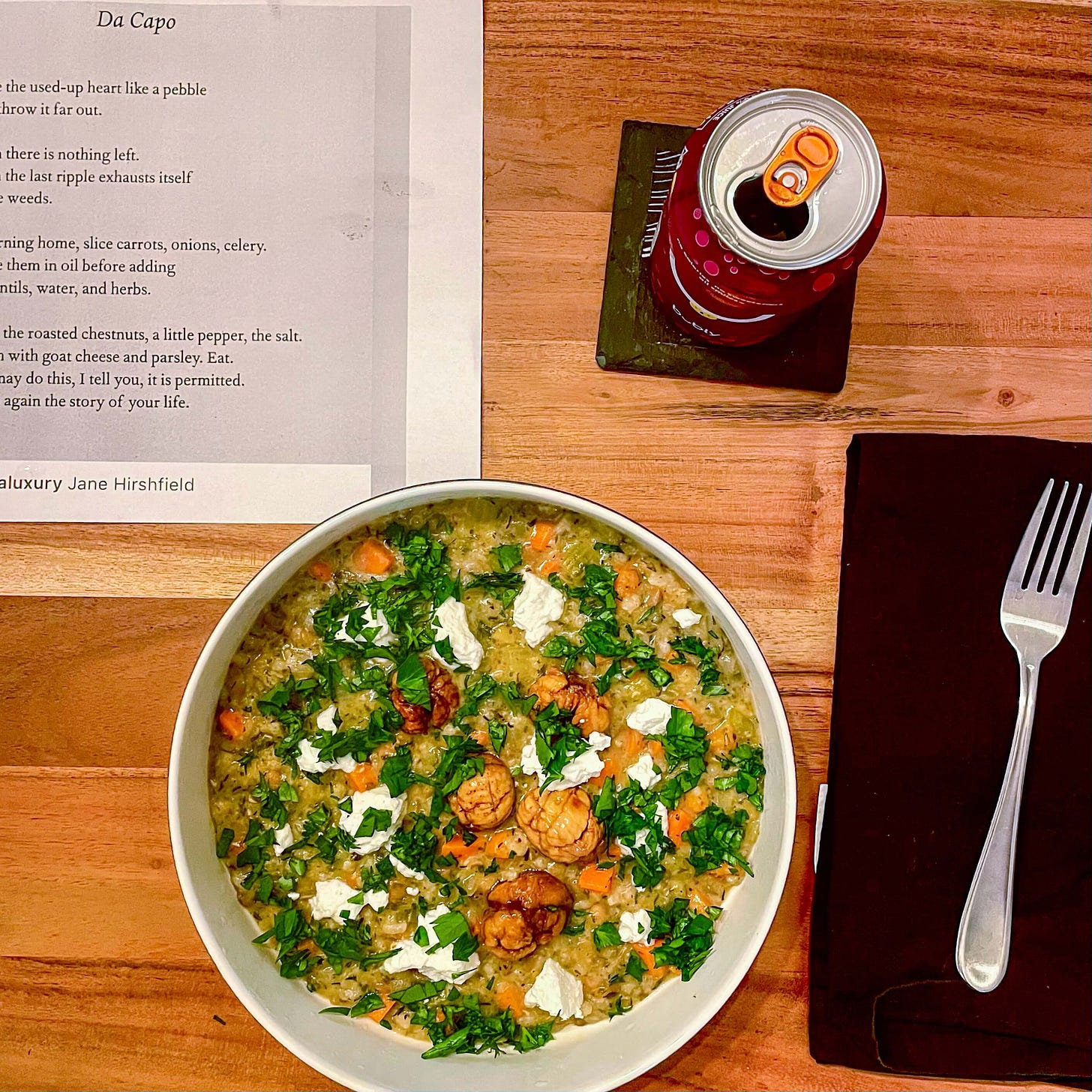 In the top left corner of the photo, the poem Da Capo by Jane Hirshfield. Next to it, a pink can of seltzer. Below, a bowl of risotto flecked with bright spots of carrots, white goat cheese crumbles, lots of parsley, and some chestnuts. To the bowl's right, a silver fork on a black napkin.