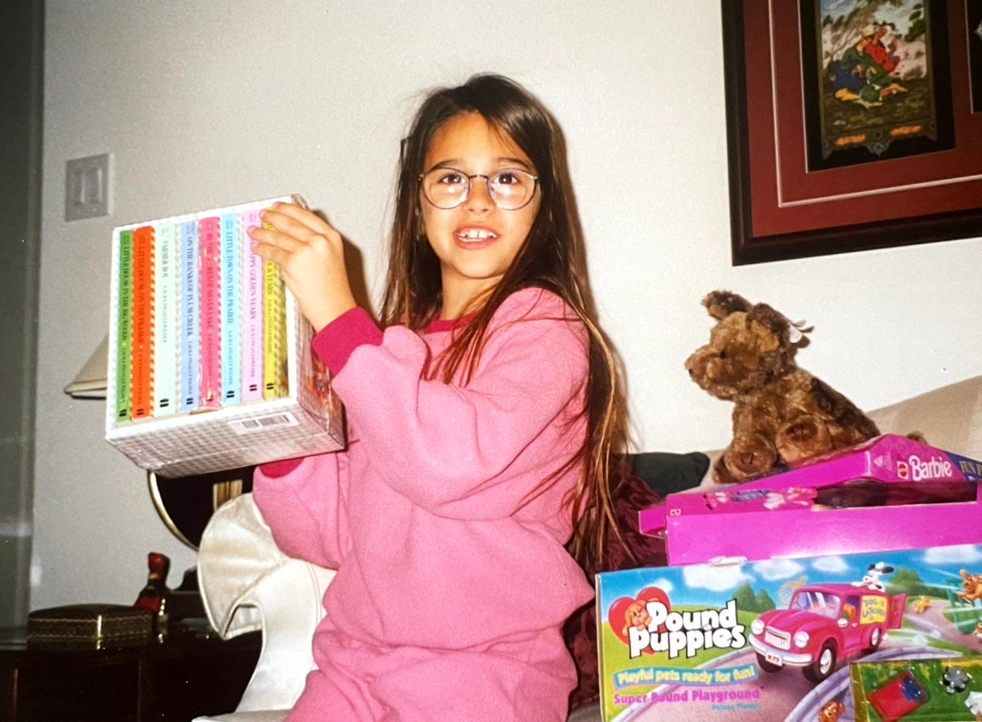 A photo of a bespectacled young Shohreh wearing a pink jogger set and holding up a box set of Boxcar Children books