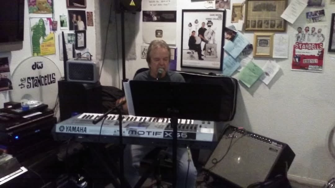 Larry Tamblyn in a home music studio. He is wearing headphones, singing into a microphone, and playing the keyboard.