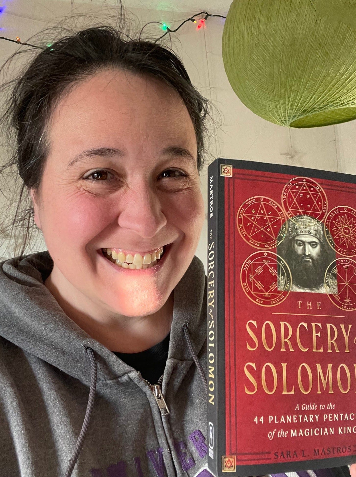 A photo of Monica holding the red and black book entitled The Sorcery of Solomon in gold capital lettering and with subtitles of “A guide to the 44 planetary pentacles of the magician king” by Sara L. Mastros.