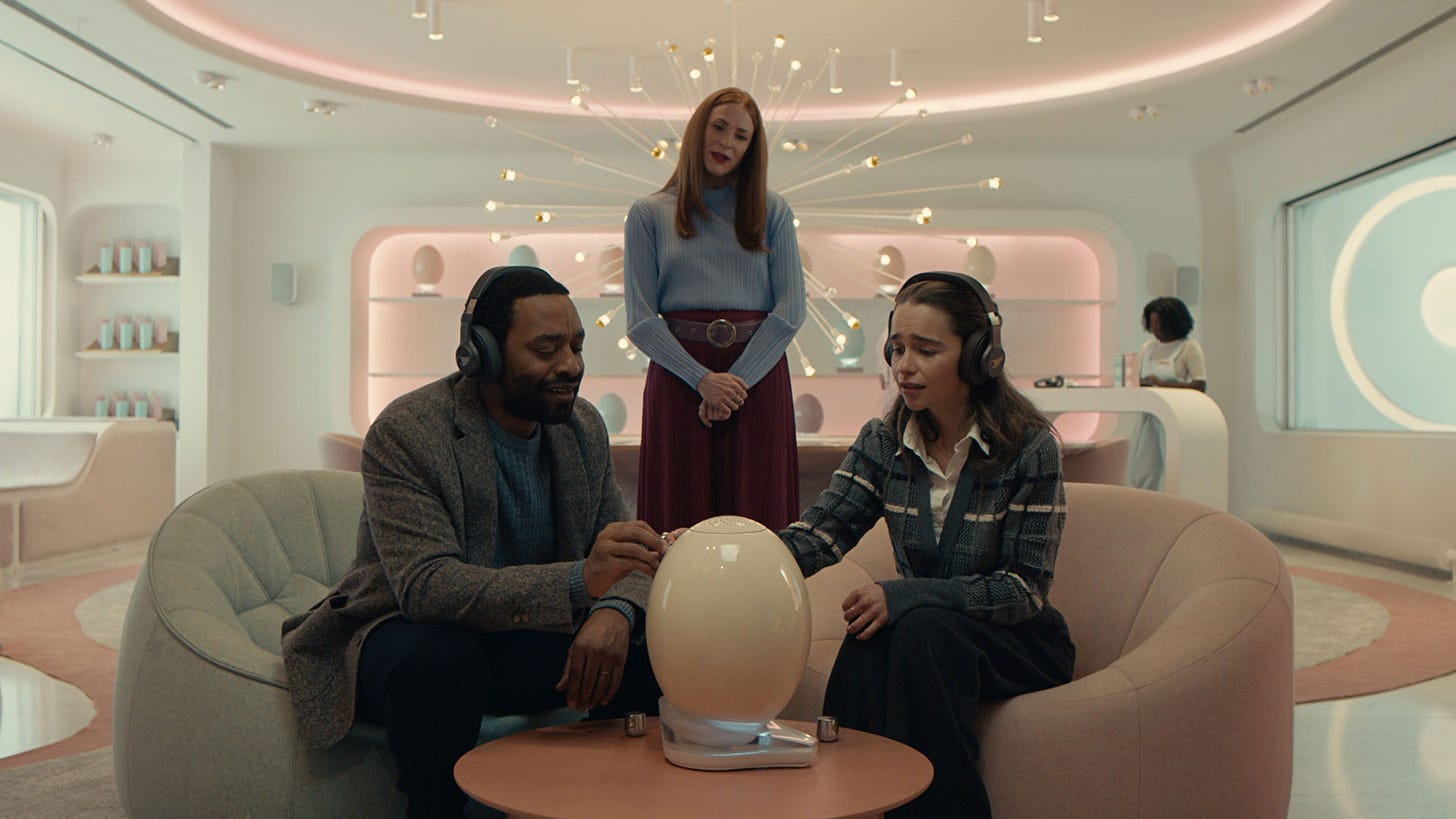 A scene from the film, The Pod Generation, where a couple have headphones on and are looking at what we assume is their fetus in an egg-like device