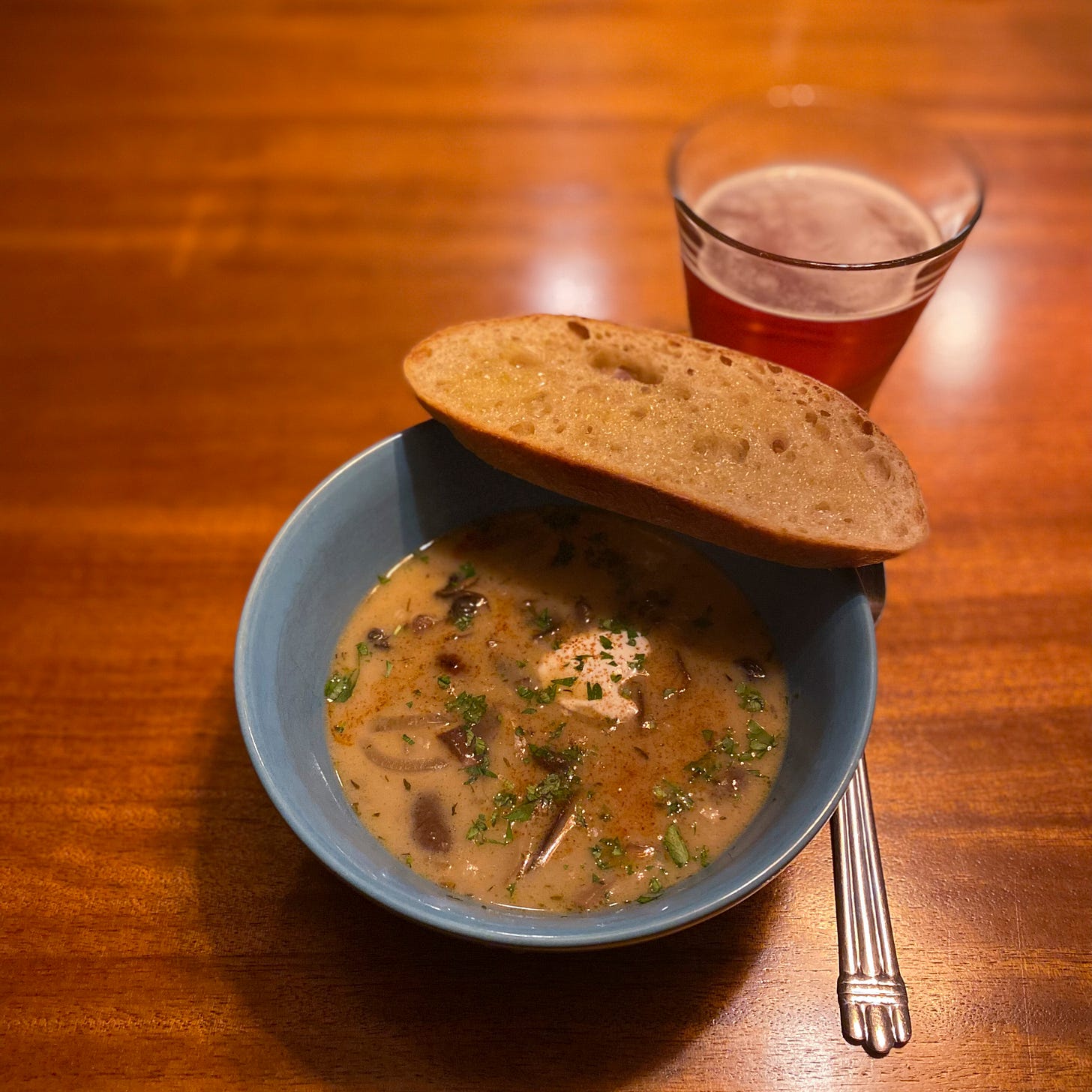 A blue bowl of creamy soup with pieces of mushroom visible throughout, and sour cream, parsley, and paprika on top. A slice of sourdough toast rests on the edge of the bowl, and a glass of amber lager sits on the table to its upper right.