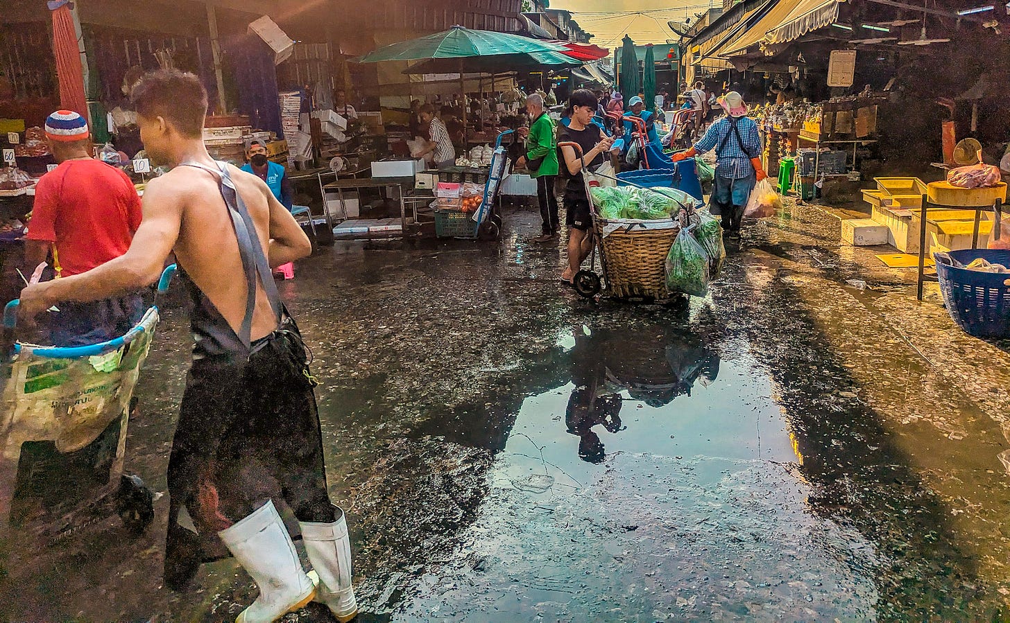 Shot of Khlong Toei market showing a young man wearing pants, an apron, and white boots pushing cart across the wet and dirty ground. 