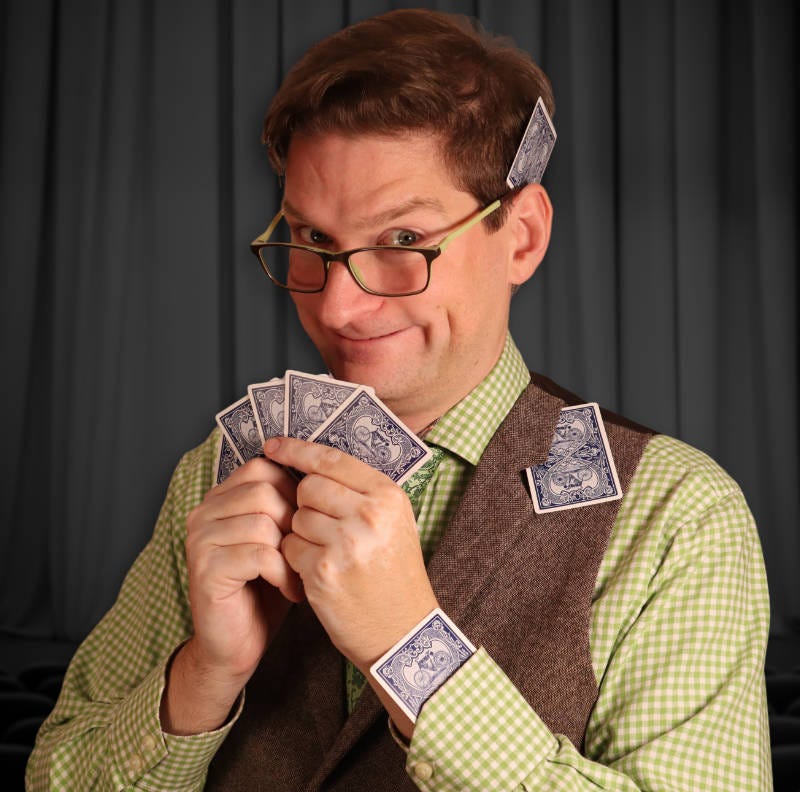 A white male with short brown hair and glasses, holding four cards. There is also a card under the lapel of his waiscoat and one up his shirt sleeve.