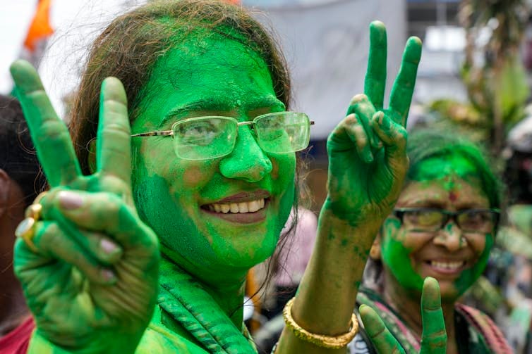 Faces of two women smeared with green color holding their fingers up in a victory sign.