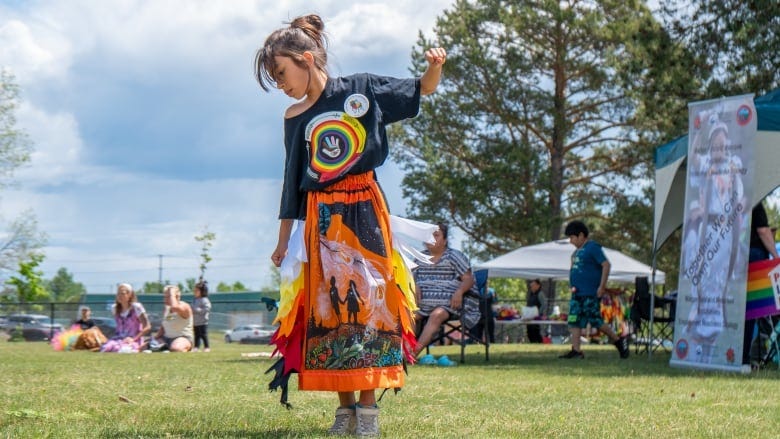 A young girl with an orange skirt is dancing on the grass with people in the background. The sky behind her is cloud. 