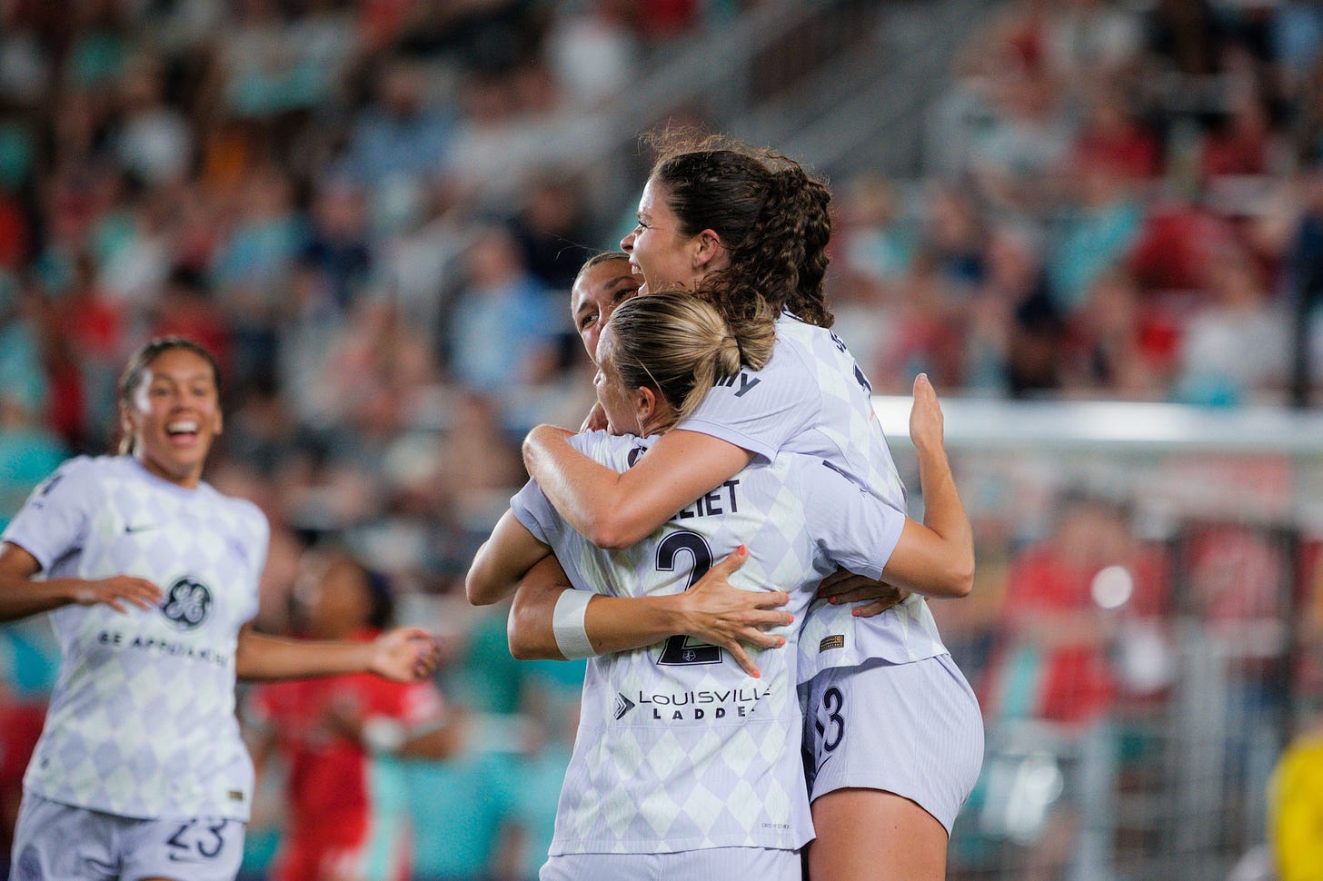 Three players in purple and white uniforms embrace while a fourth player runs up to them smiling