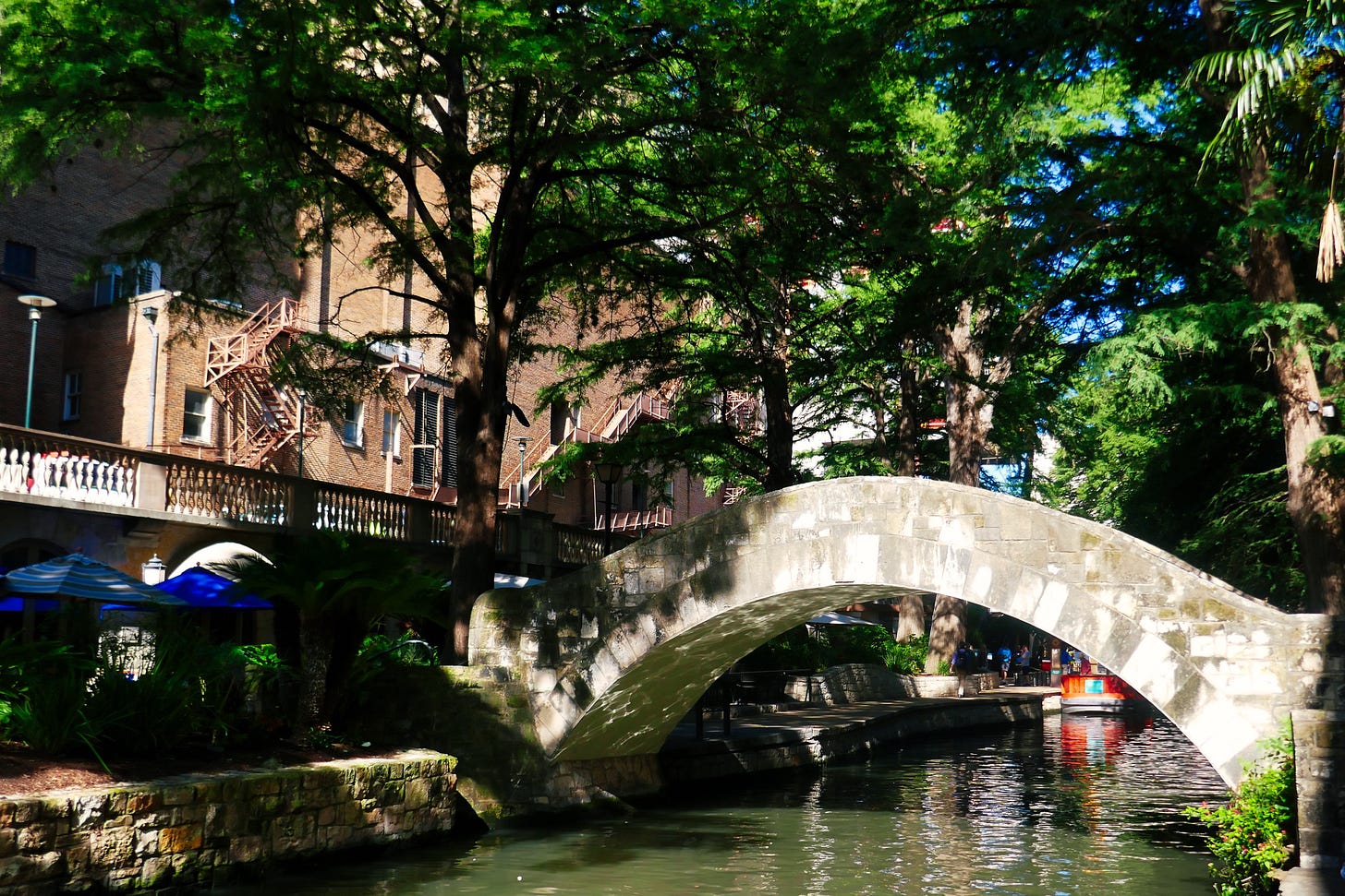 A footbridge across the narrow San Antonio River under trees