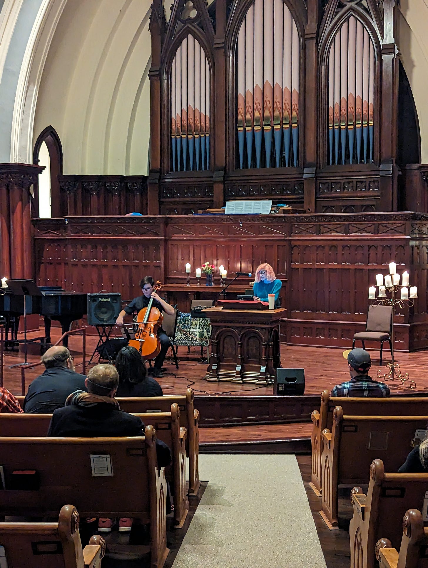 Elle sitting behind pulpit and Julian holding their cello on the "stage" in Sanctuary Hall with the gran organ behind them and candelabras flanking them with audience members in view