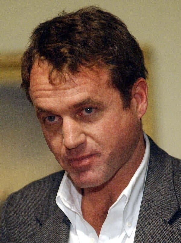 A close-up portrait. He has brown hair and wore a gray sport jacket over an open-collared white shirt.