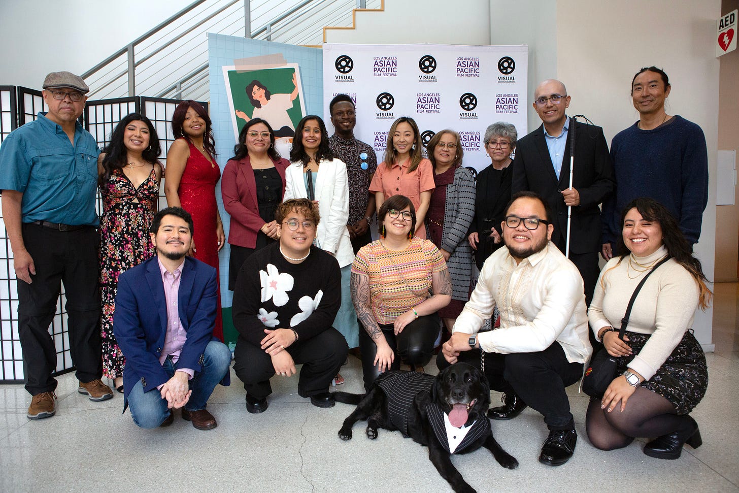  A multiracial and intergenerational group of people pose for the camera in two rows in front of signage for a film festival. 11 people on the back row are standing. 5 people (and a black Labrador) squat on the front row.