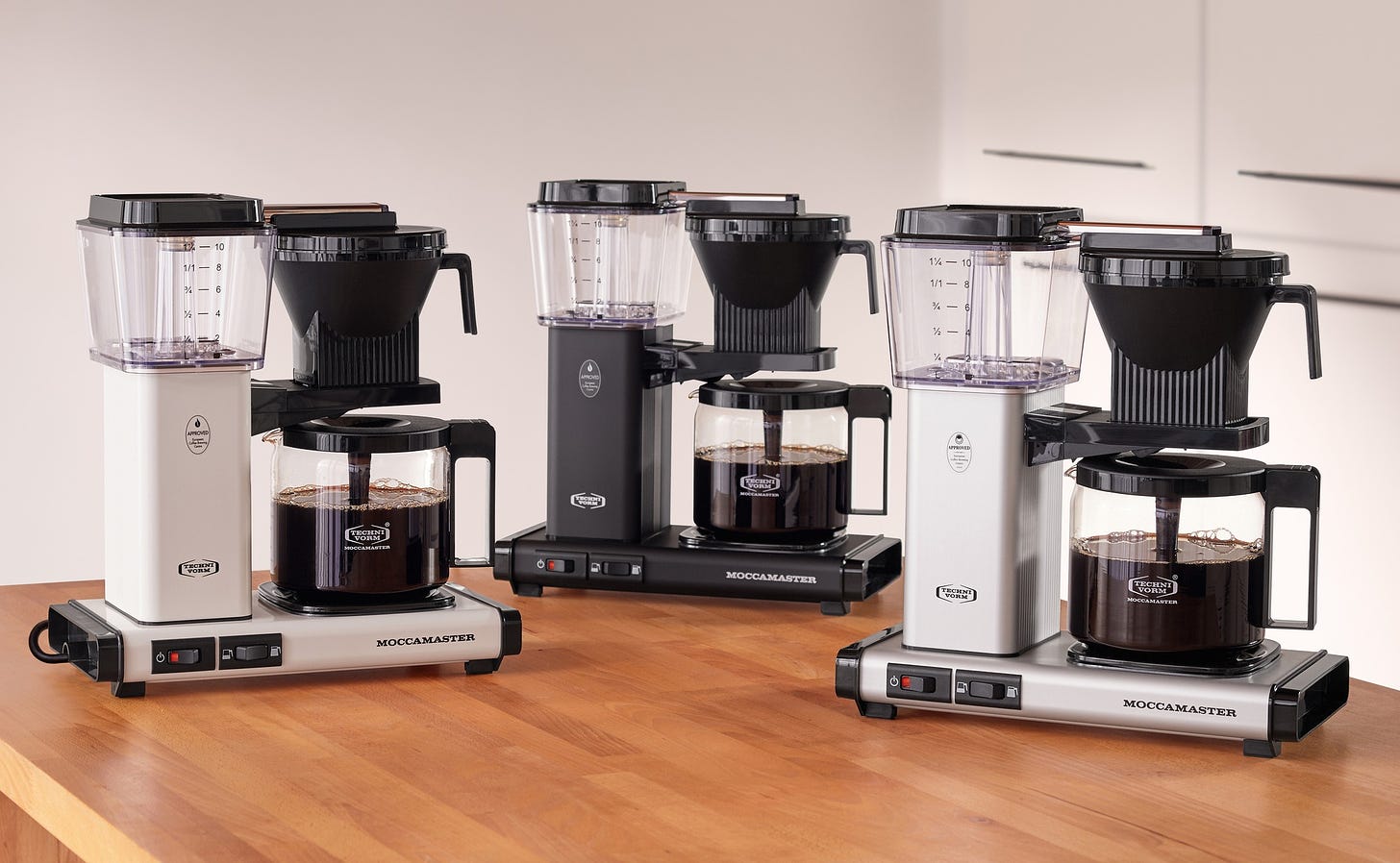 Three automatic coffee brewers lined up on a wood tabletop.