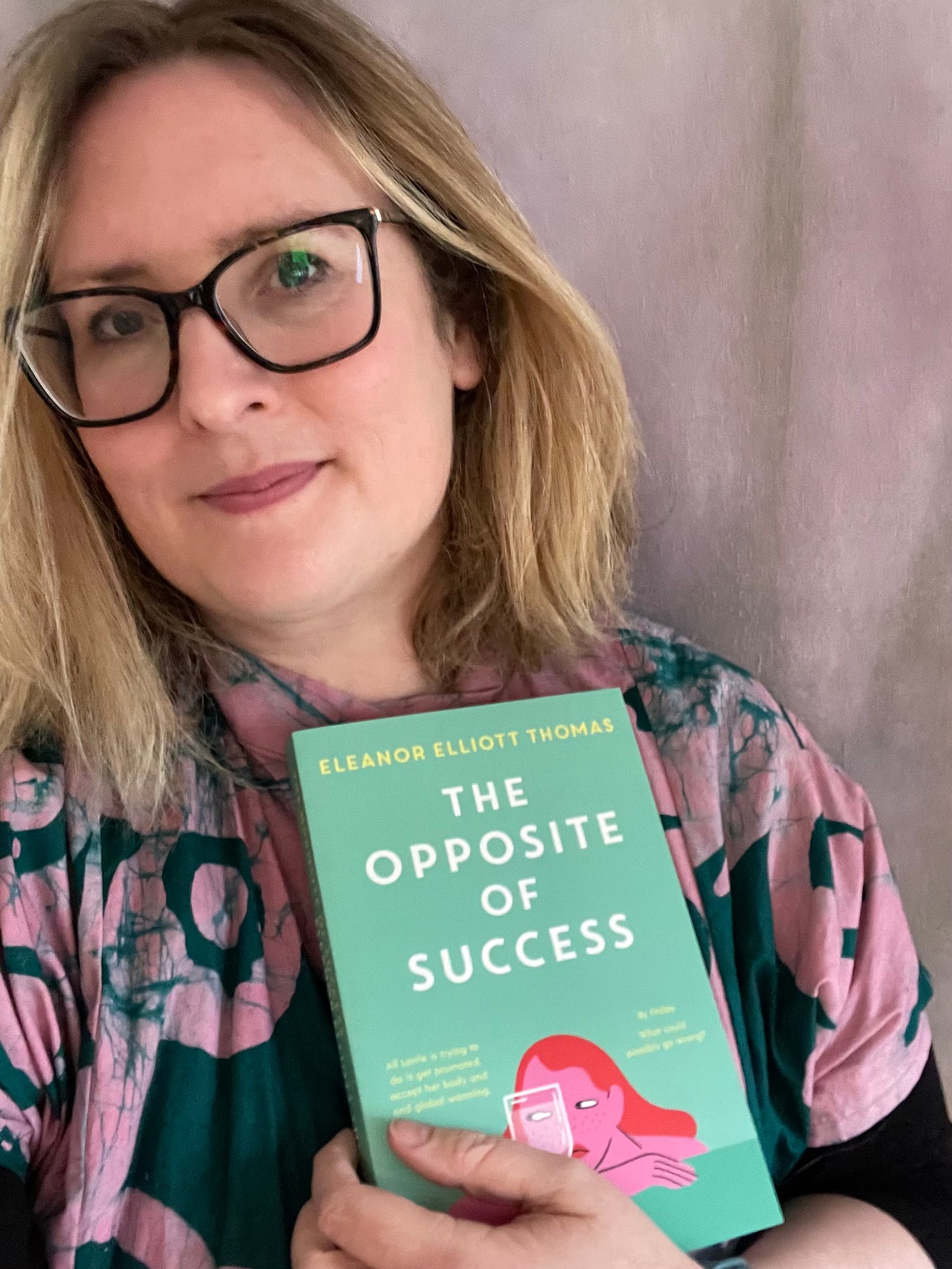 The author, a blonde woman in glasses and a pink and green top, looks at the camera while holding a copy of The Opposite of Success, which has a green cover with a cartoonish illustration of a woman looking sideways