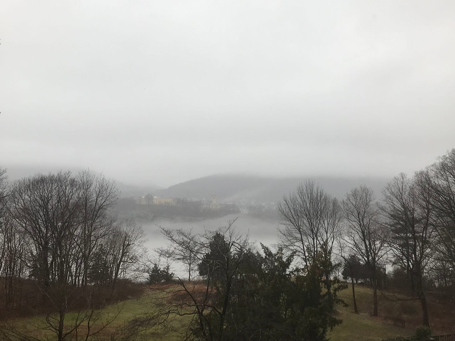 A view of the Hudson river half-obscured by mists. In the foreground are bare winter trees, and across the river are blurred hills.