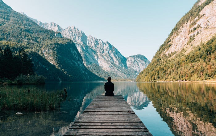 A person sits alone at the end of a wooden walkway that leads out from the shoreline into a lake. The lake is surrounded by mountains. The sky is blue and the lake is still reflecting the scenery around it.
