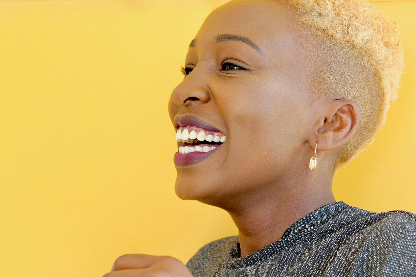 This image features a close-up of a smiling person with short, blonde hair against a bright yellow background. The individual is shown in profile, with a wide smile that reveals their teeth. They are wearing a small hoop earring and a textured, dark-colored top. The overall tone of the image is vibrant and cheerful.