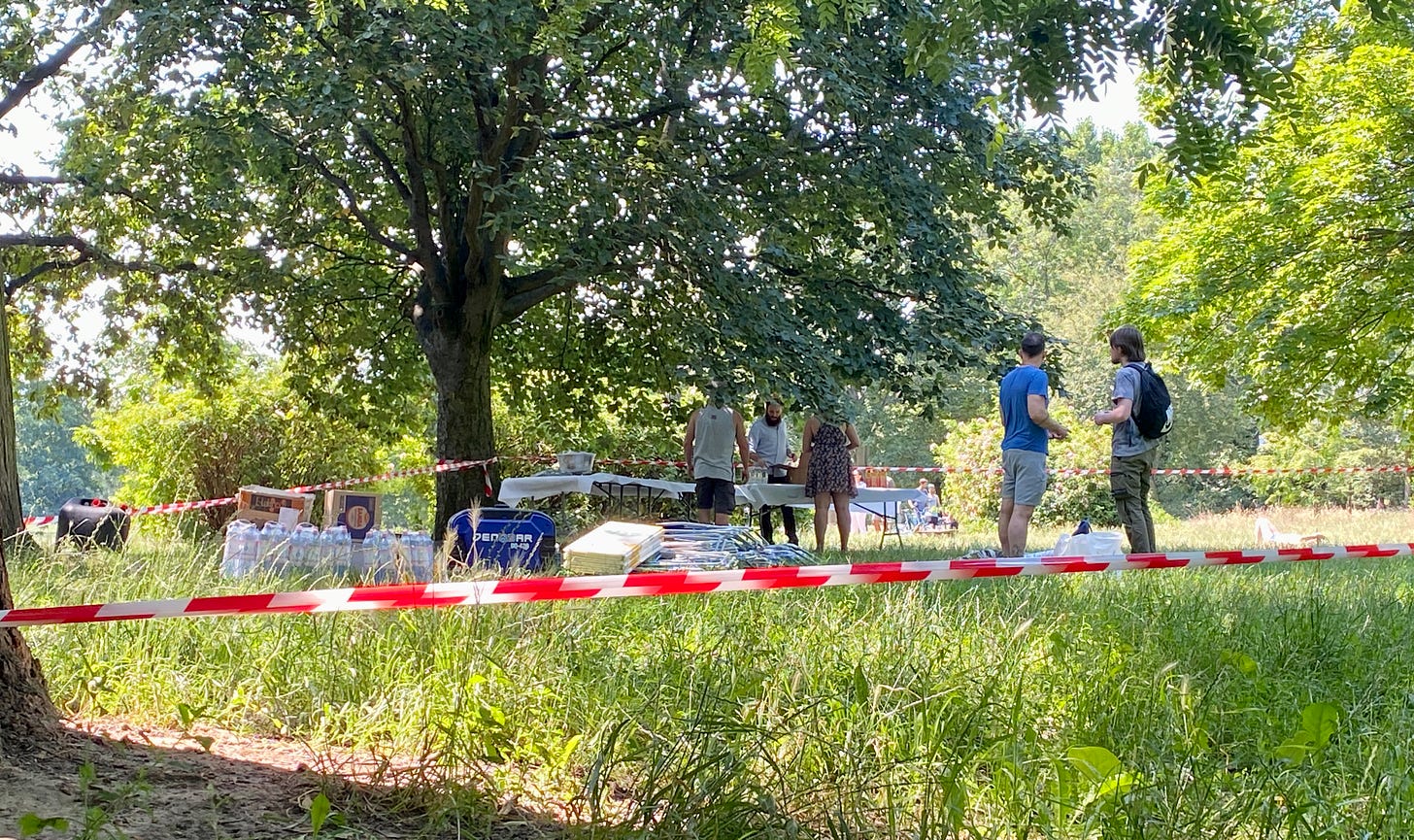 Imagem: gramado verde, arvores, uma faixa branca e vermelha cercando tudo, no meio 2 homens conversam, tem várias caixas e pessoas montam mesas nos fundos