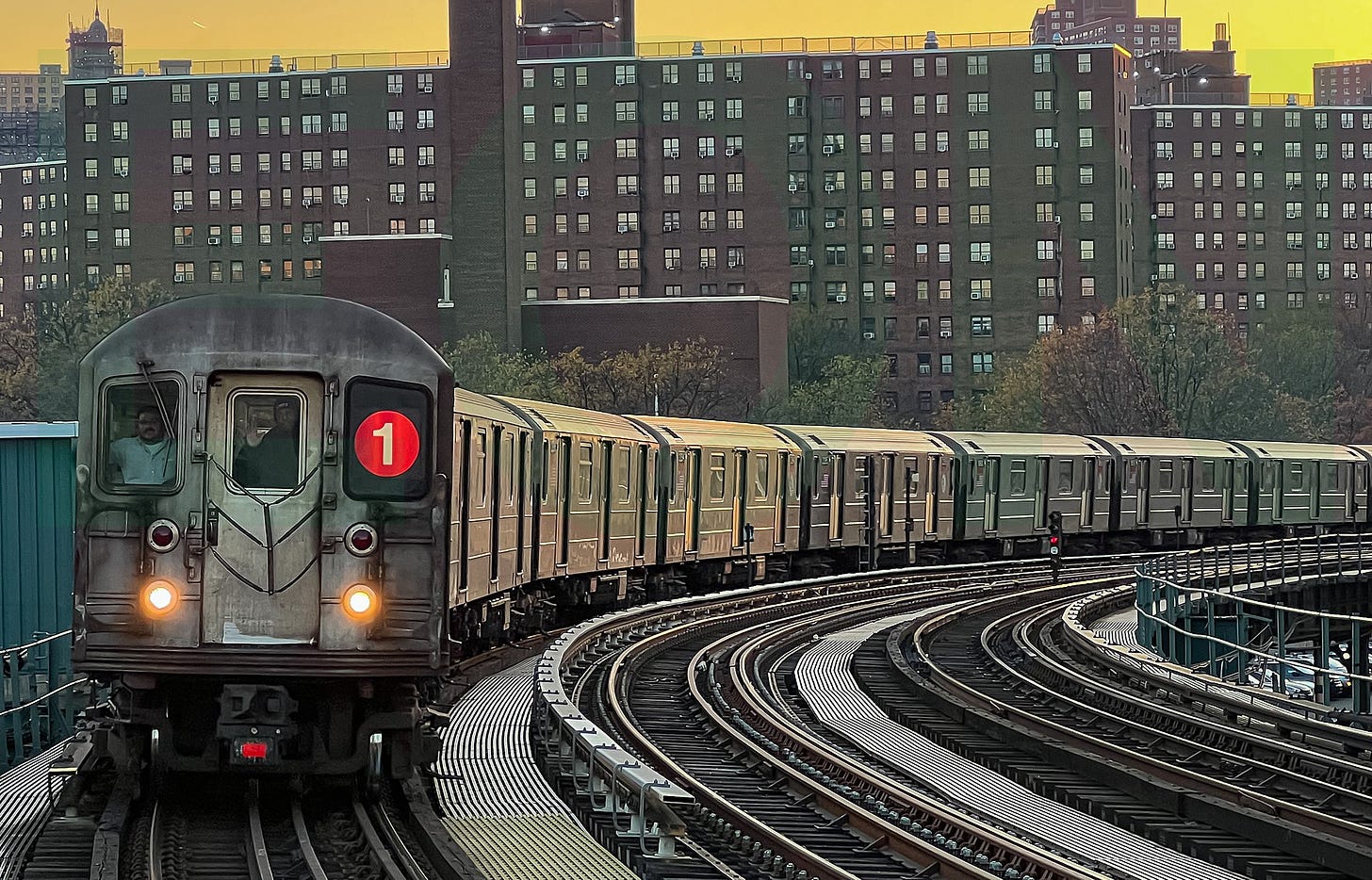 A 1 train, composed here of R62A cars is seen above ground entering the 207th Street station. The front of the train contains two white lights providing slight illumination, two windows, a door, and the Symbol for the 1 line on the left window.