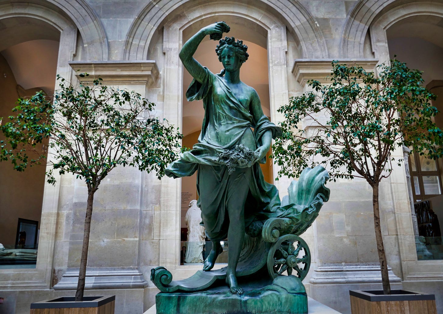 A bronze statue in the Louvre in Paris of a woman holding pinecones and stepping from a small chariot, two small trees flank her on either side