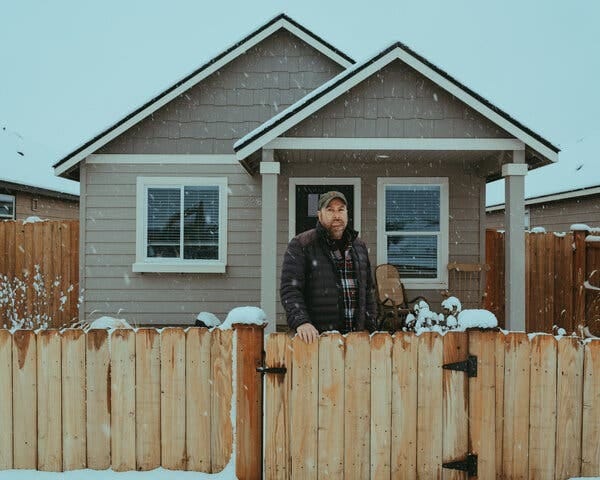 Robert Lanter standing behind a waist-high wooden fence, with his small home in the background.