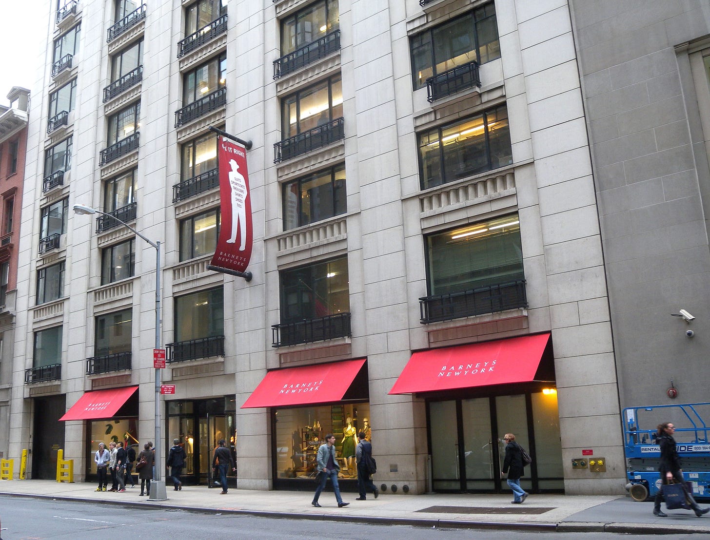 A view of the Barneys New York flagship exterior with bright red awnings over the first floor windows that read, "Barneys New York". Everyday New Yorkers walk past the building facade glancing into its display windows.