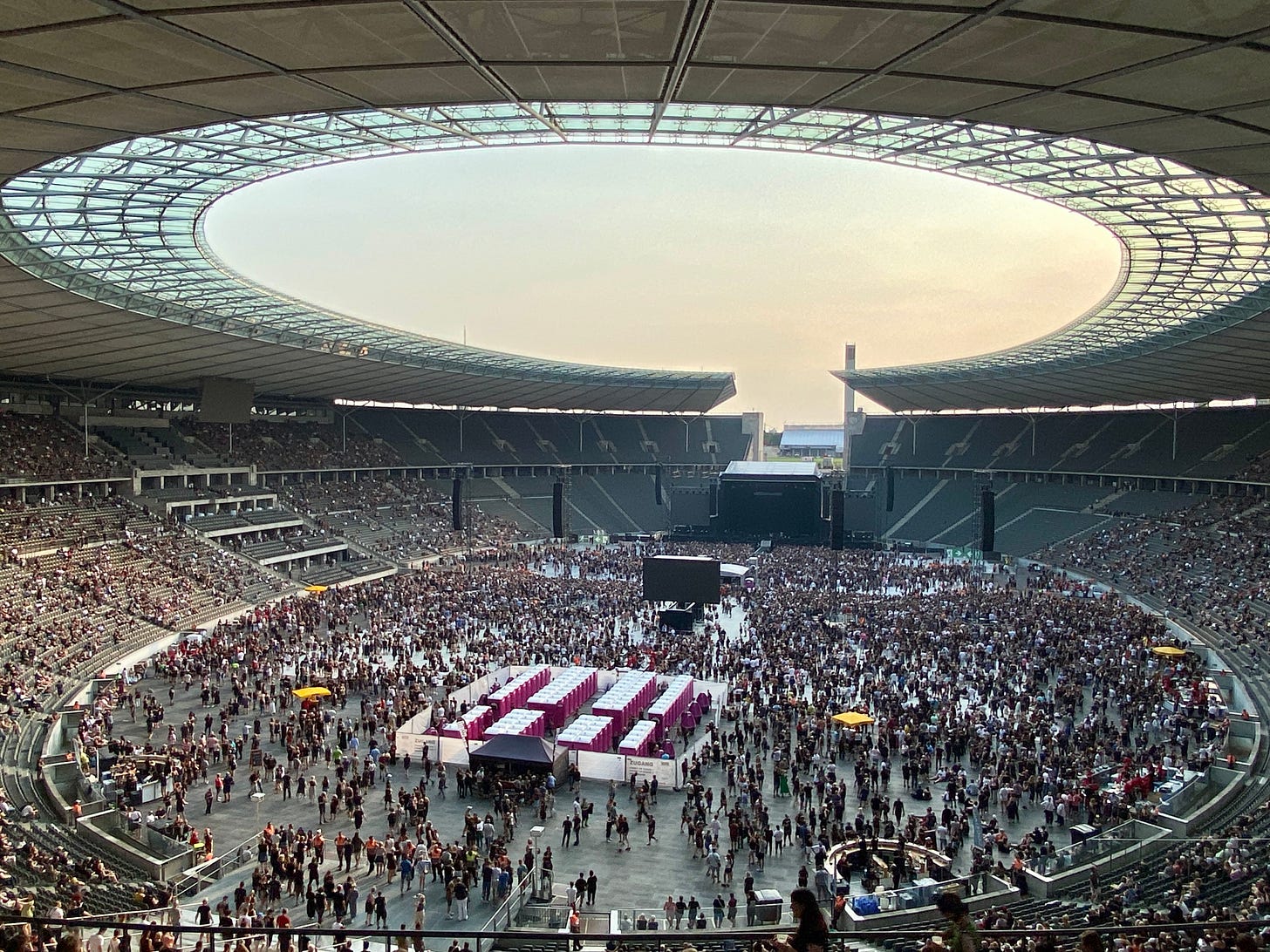 Imagem do estádio vista de cima. Teto oval aberto com o céu laranja. A pista está lotada de gente, tem um telão no meio e o palco aos fundos.